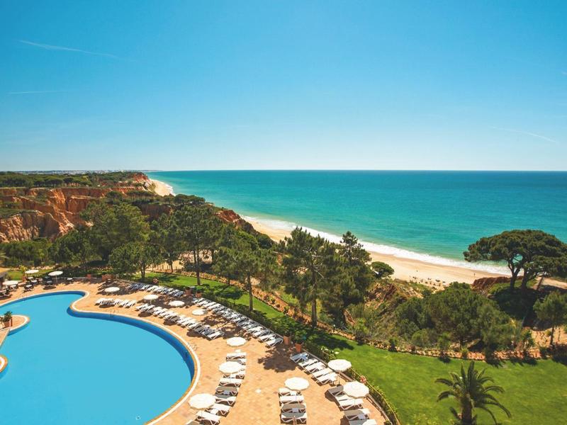 Hotelový bazén s lehátky, palmami a výhledem na dlouhou pláž a modré moře.