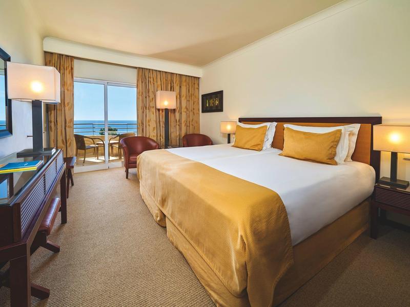 Světý hotelový pokoj s velkou postelí, stolem, křeslem a výhledem na moře.