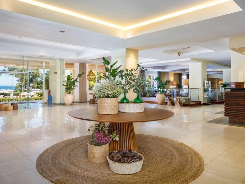 Světlá hotelová hala s kulatými stoly, rostlinami a útulnými posezeními.