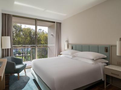 Moderna habitación de hotel con gran cama, sillón, televisor y balcón con vista a áreas verdes.