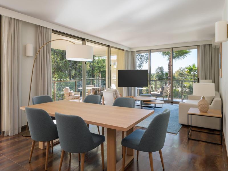 Área de estar y comedor luminosa con mesa de madera, sillas modernas y gran ventanal al balcón con plantas.