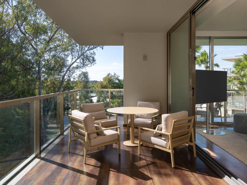 Balcón con mesa y cuatro sillas, rodeado de barandilla de vidrio y vista a árboles y habitación adyacente.