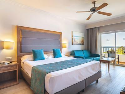 Lichte hotelkamer met groot bed, turkooizen accenten, bank, balkon en plafondventilator.