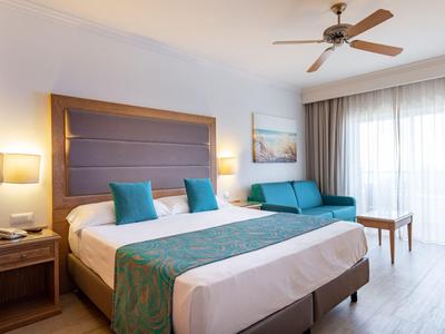 Helles Hotelzimmer mit Doppelbett, blauen Akzenten und Balkon mit Vorhängen.