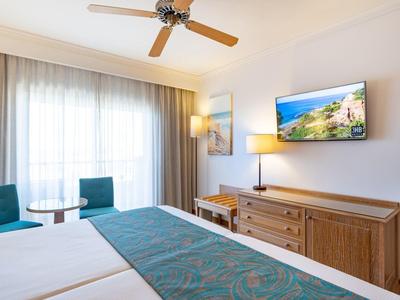 Lichte hotelkamer met twee eenpersoonsbedden, turquoise accenten, houten meubels, plafondventilator en balkonlicht.