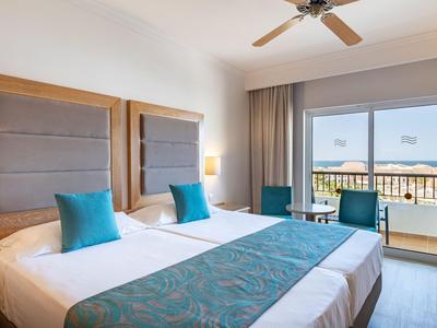 Helles Hotelzimmer mit zwei Betten, blauen Akzenten und Balkon mit Meerblick.