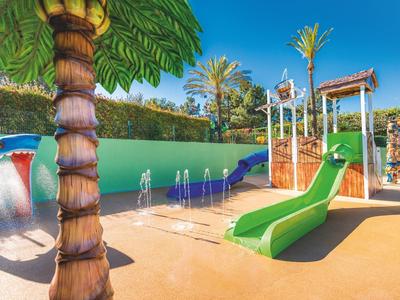 Zonnige buitenwaterspeeltuin met glijbanen, fonteinen en palmboomdecoraties.