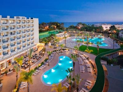 Resorthotel bij zonsondergang met zwembaden, palmbomen en ligstoelen aan zee.