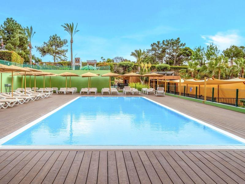 Buitenzwembad rechthoekig met ligstoelen en groen onder een blauwe lucht.