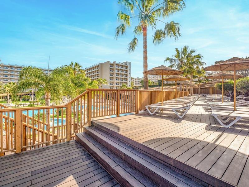 Ruim zwembadterras met ligstoelen, palmbomen en een zonnige blauwe lucht bij een resorthotel.