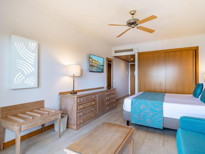 Lichte hotelkamer met blauwe accenten, houten meubels, plafondventilator, bed en tv.