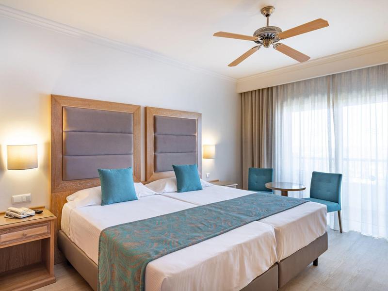 Moderne hotelkamer met twee eenpersoonsbedden, teal accenten, houten meubels en doorschijnende gordijnen.