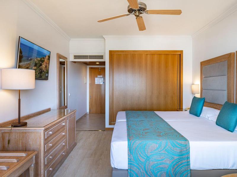 Lichte hotelkamer met twee bedden, blauwe accenten, houten meubels en een plafondventilator.