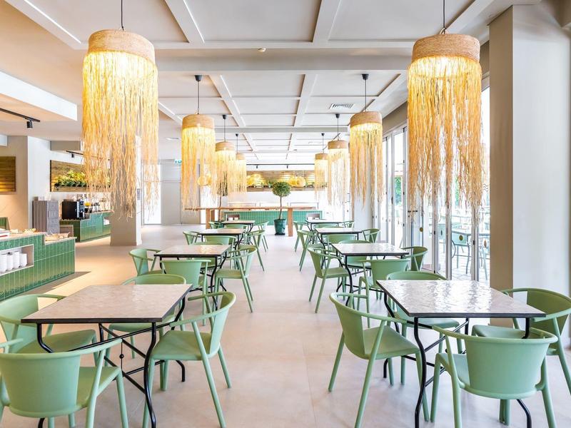 Moderne eetruimte met groene stoelen, beige tafels en grote hanglampen van geweven materiaal.