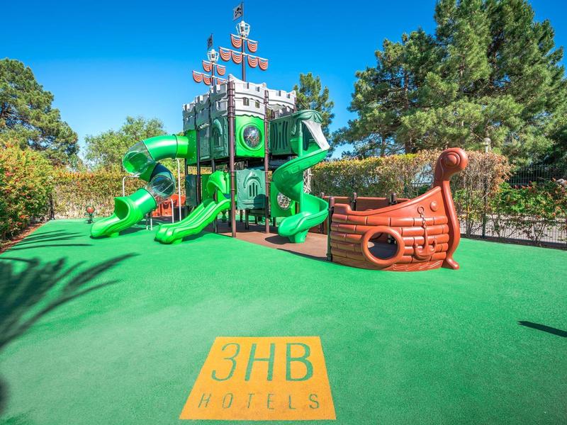 Buitenspeeltuin met piratenschip en glijbanen op groene rubberen vloer onder blauwe lucht.