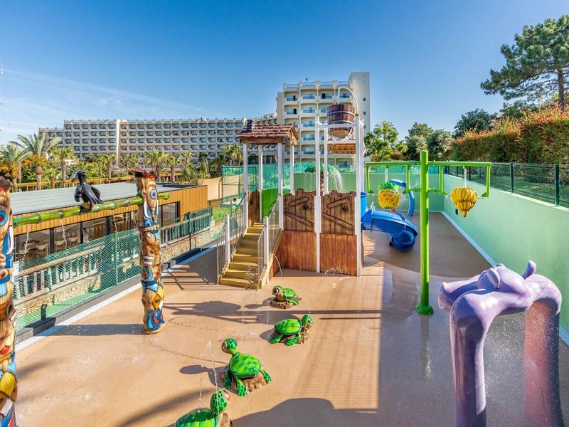 Buitenspeeltuin met glijbanen en dierenvormige wipkippen naast hotelgebouw onder blauwe lucht.