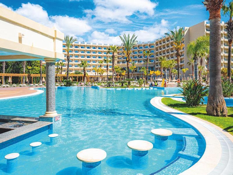 Resortzwembad met ronde opstapkrukken en palmbomen eromheen onder een blauwe lucht.