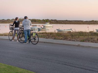 Zwei Männer mit Fahrrädern stehen an einer Straße und blicken auf Boote im Wasser bei Sonnenuntergang.
