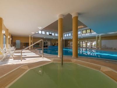 Hallenbad mit großem Schwimmbecken, Whirlpool, gelben Säulen und Sonnenliegen am Rand.