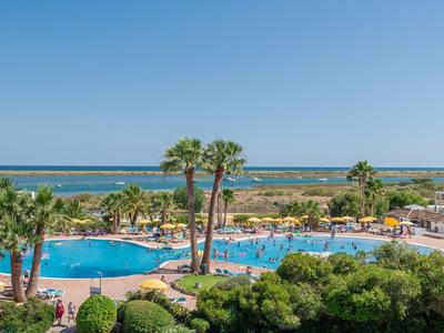 Großer blauer Pool umgeben von Palmen und grünen Pflanzen, im Hintergrund das blaue Meer.