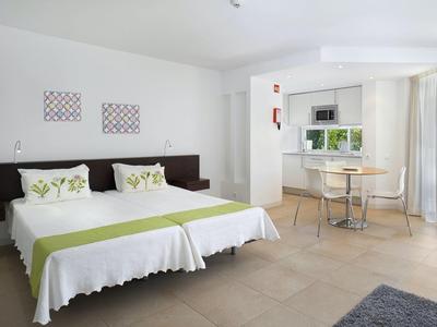 Helles Zimmer mit zwei Betten, grünen Akzenten, rundem Tisch, Stühlen und großen Fenstern.