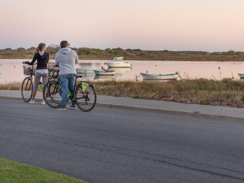 Zwei Männer mit Fahrrädern stehen an einer Straße und blicken auf Boote im Wasser bei Sonnenuntergang.