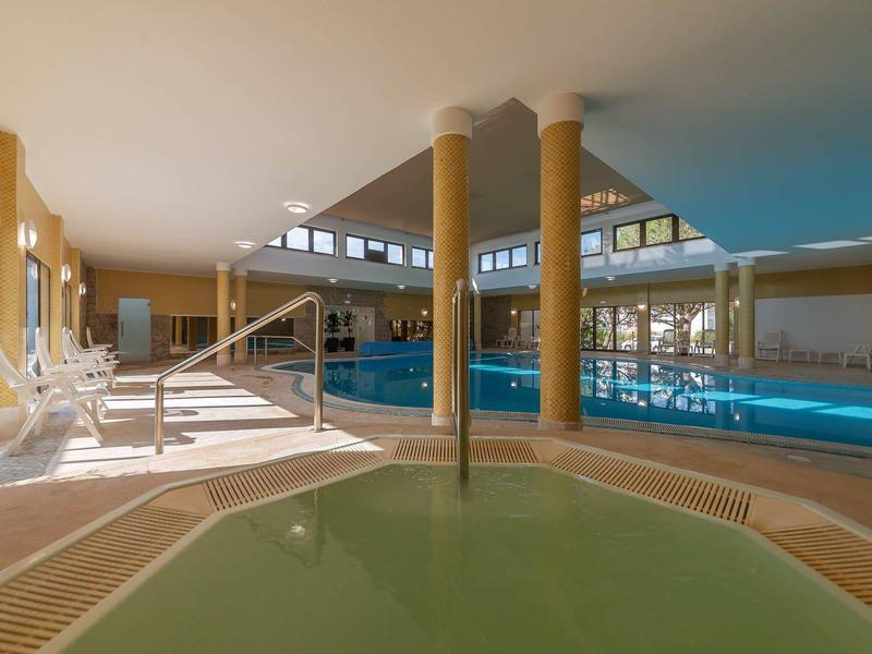 Hallenbad mit großem Schwimmbecken, Whirlpool, gelben Säulen und Sonnenliegen am Rand.