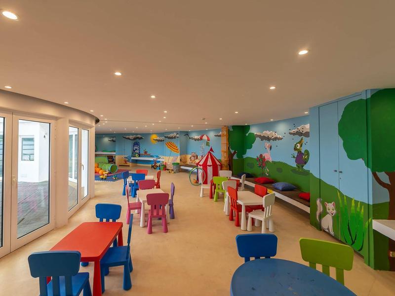 Buntes Kinderzimmer mit kleinen Tischen, Stühlen und Wandmalereien von Natur und Spielzeug.