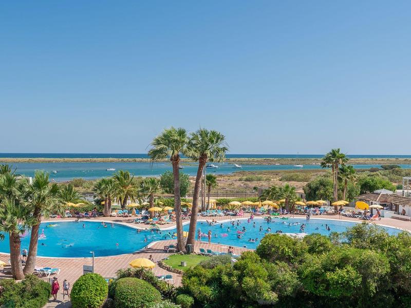 Großer blauer Pool umgeben von Palmen und grünen Pflanzen, im Hintergrund das blaue Meer.