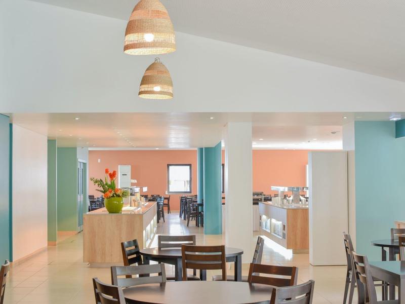 Moderner Gastraum mit Holztischen, Stühlen, blauen Wänden und orangefarbenem Hintergrund.