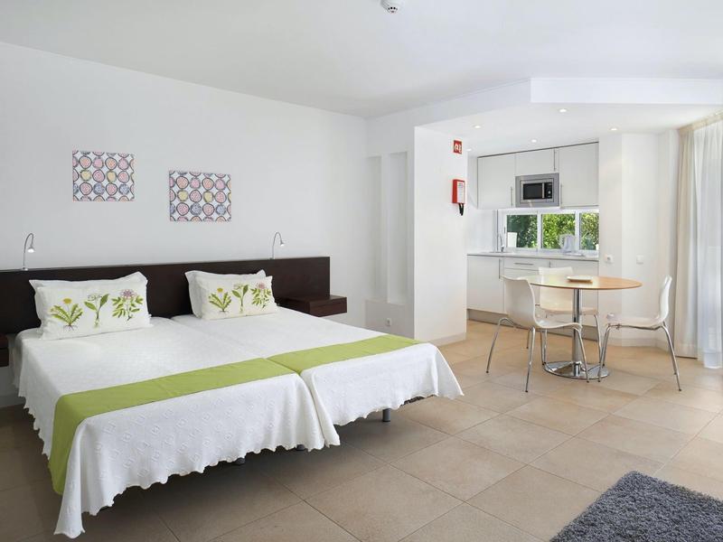 Helles Zimmer mit zwei Betten, grünen Akzenten, rundem Tisch, Stühlen und großen Fenstern.