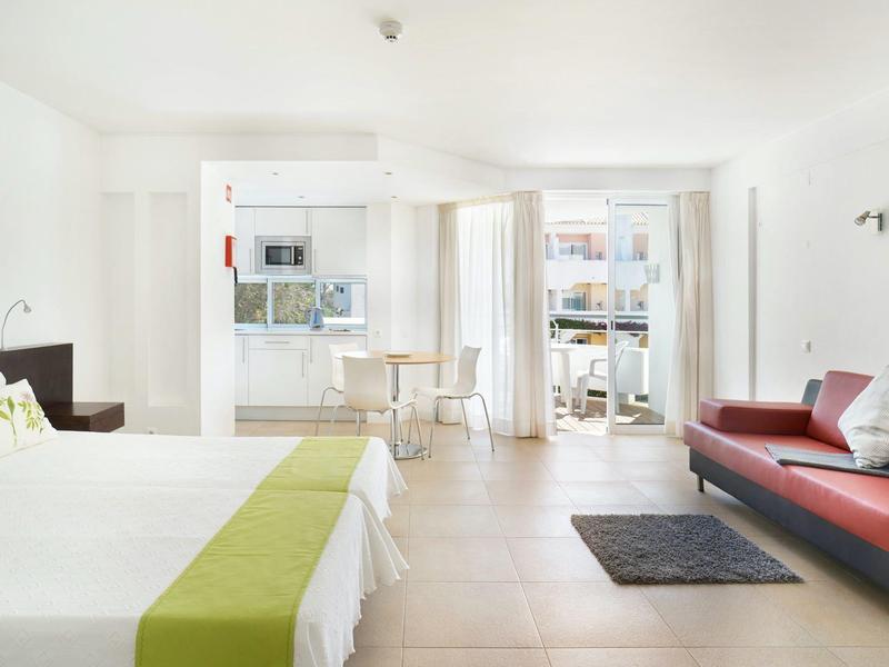 Helles Zimmer mit Doppelbett, rotem Sofa, kleinem Esstisch und Balkontür mit Vorhängen.