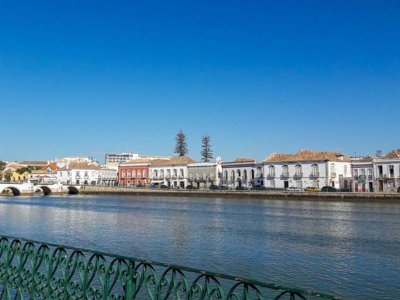 Flussufer mit weißen Häusern, klarem Himmel und grünem Geländer im Vordergrund.