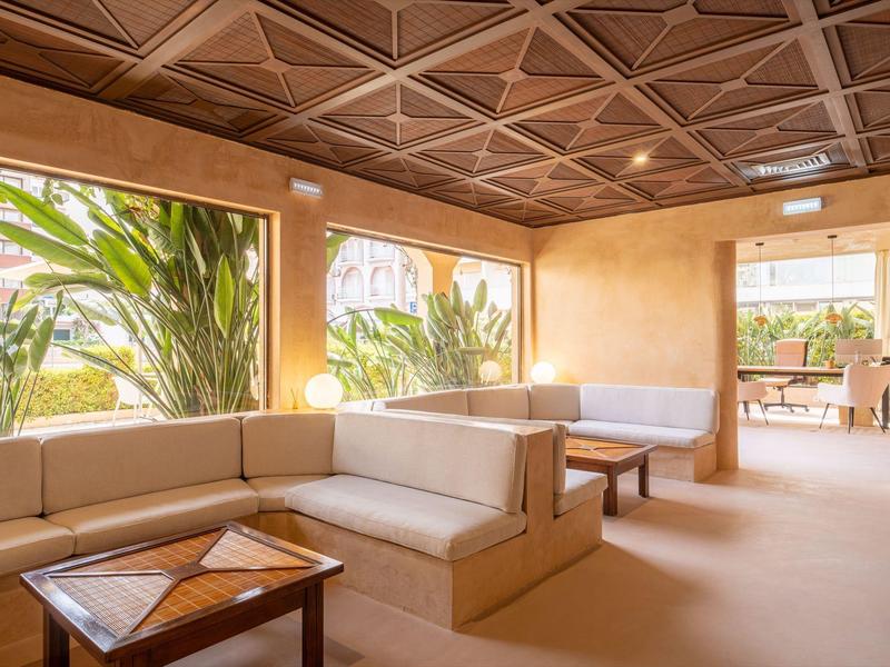 Bright lounge with white sofas, wooden tables, and large windows overlooking tropical plants.