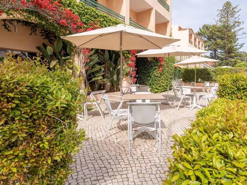 Terrace with tables, chairs, and umbrellas in front of a greenery-covered building on a sunny day.