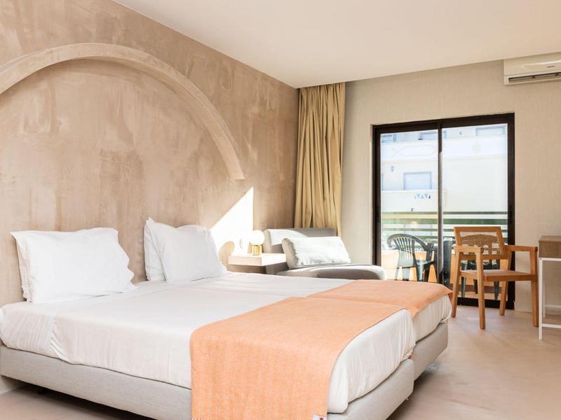 Bright hotel room with two beds, beige walls, and terrace overlooking the sea.