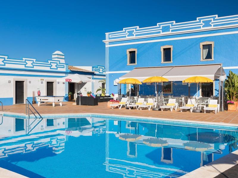 Hotel met blauw geschilderd gebouw en groot buitenzwembad onder heldere lucht.