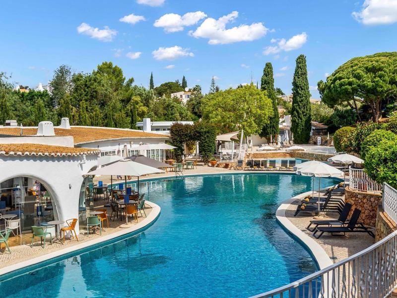 Zwembad met ligstoelen en parasols naast een hotel onder een blauwe lucht.