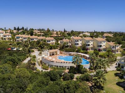 Ośrodek wypoczynkowy z willami i okrągłym basenem otoczonym zielenią pod niebieskim niebem.