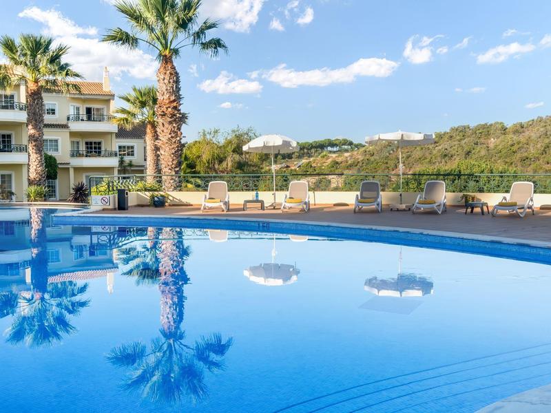 Okrągły basen hotelowy z palmami i leżakami na tle gór pod błękitnym niebem.