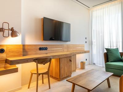 Jasny pokój hotelowy z telewizorem na ścianie, biurkiem, krzesłem, zieloną sofą i dużym oknem z zasłonami.