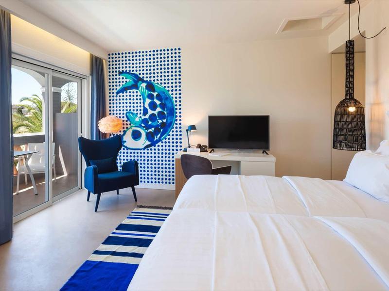 Moderne hotelkamer met tweepersoonsbed, blauwe fauteuil en balkon met zeezicht.