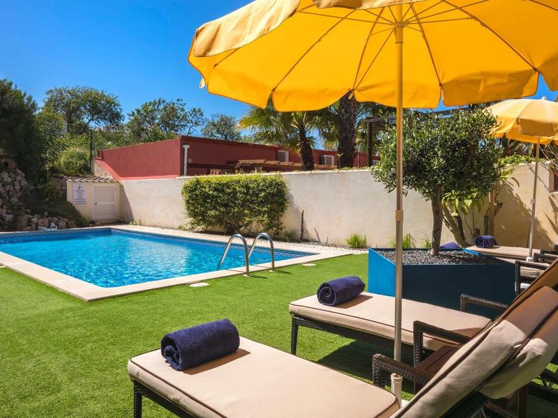 Zonnig zwembad met ligstoelen en gele parasols in een hoteltuin.