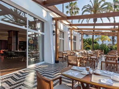 Open eetruimte met houten tafels en witte stoelen onder een pergola naast een restaurant.