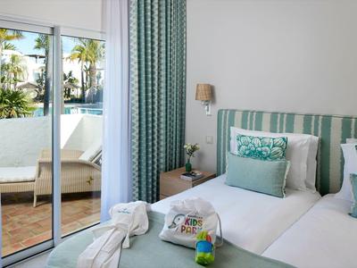 Lichte hotelkamer met twee bedden, turquoise kussens en balkon met uitzicht op het zwembad.