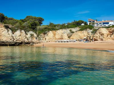 Helder zeewater bij een rustig zandstrand met rotsen en huizen op de achtergrond.