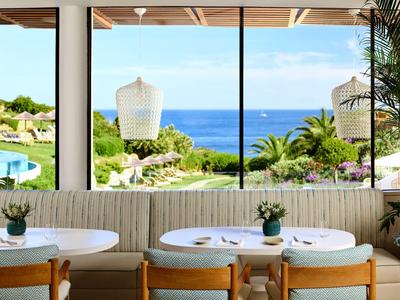 Moderner Essbereich mit Holzstühlen, weißen Tischen und großem Fenster mit Blick auf Meer und Garten.