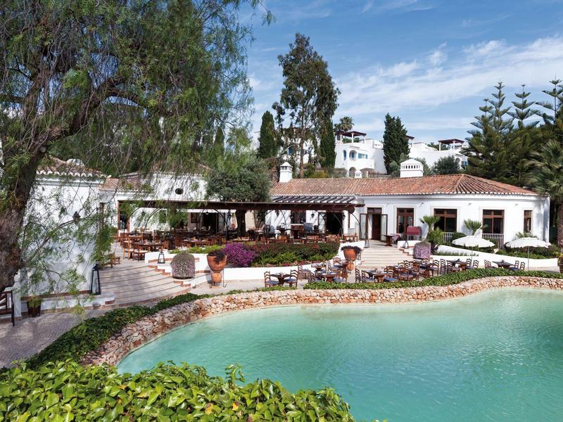 Hotelgebouw met terras en zitplaatsen bij een blauw zwembad omringd door bomen.