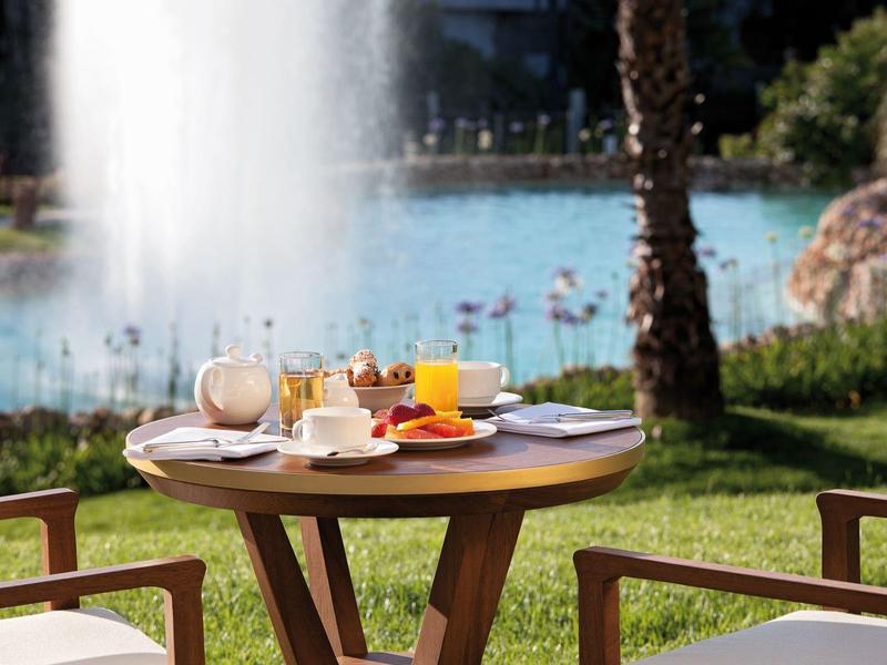 Kleine ronde houten tafel met ontbijt bij fontein en meer op de achtergrond.