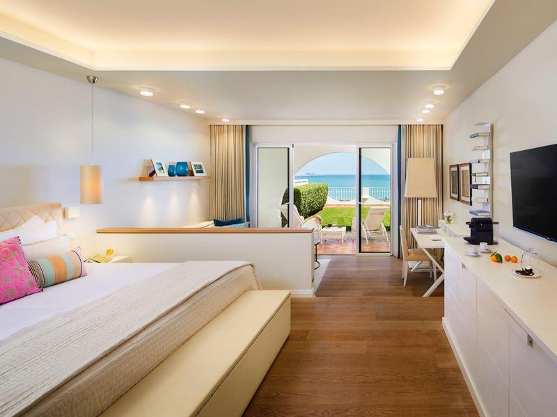 Lichte hotelkamer met tweepersoonsbed, tv, bureau en uitzicht op zee en tuin.